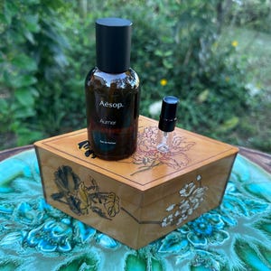 May include: A brown glass perfume bottle with a black cap and the text "Aesop. Aumer eau de parfum" sits on a wooden box with floral designs. A small glass vial with a black cap is next to the bottle.