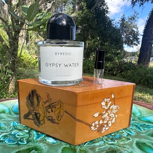 May include: A clear glass perfume bottle with a black cap and the text "BYREDO GYPSY WATER" sits atop a wooden box with floral designs. A small perfume sample bottle is next to it. The box is on a green and blue decorative plate.