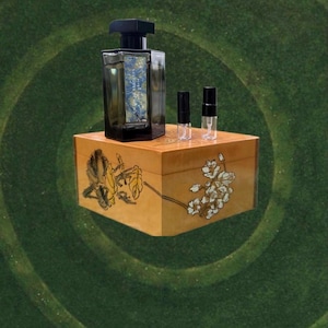 May include: A dark-colored perfume bottle with a black cap and a decorative label, accompanied by two small sample vials, rests on a wooden box. The box is adorned with floral artwork in yellow and white, set against a green background.