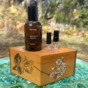 May include: A brown glass bottle of Aesop Marrakech Intense Eau de Parfum with a black cap, and two small sample vials, sit atop a decorative wooden box with floral designs. The box is on a green and blue decorative plate.