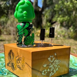 May include: A green-topped perfume bottle with a whimsical design, set atop a decorative wooden box with floral artwork. Two small perfume vials sit beside the bottle. The box is on a green and blue surface.