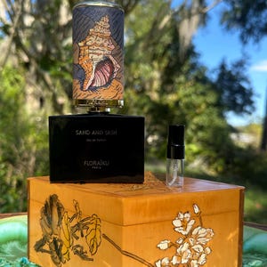 May include: A black rectangular perfume bottle with the words "SAND AND SKIN" and "FLORAIKU PARIS" printed on it. A small glass vial and a decorative perfume bottle with a seashell design sit on a wooden box with floral artwork.