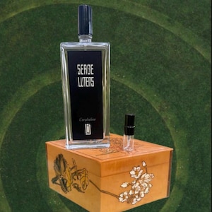 May include: A rectangular perfume bottle with a black label that reads "SERGE LUTENS" and "L'orpheline". A small sample bottle sits next to it on a wooden box with floral designs. The bottle has a black cap.