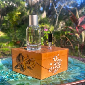 May include: A clear glass perfume bottle with a silver cap and a smaller sample bottle sit atop a decorative wooden box. The box is orange with floral designs. The background is a blurred outdoor scene.