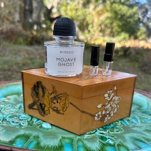 May include: A clear glass perfume bottle with a black cap and the text "BYREDO MOJAVE GHOST" sits atop a small, decorated wooden box. Two small vials are next to the perfume bottle. The box is on a green and blue decorative plate.