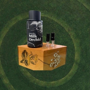 May include: A black perfume bottle with the text "commodity Milk Orchid" and two small sample vials sit atop a decorative wooden box. The box is painted with floral designs in gold and white.