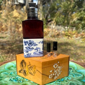 May include: A perfume bottle with a dark red body and a silver cap sits atop a wooden box. The bottle has a white base with blue bird and floral designs. Two small sample vials are next to the bottle. The box has floral designs.