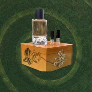 May include: A perfume bottle labeled "NO 1" with a black cap, sitting on a wooden box with floral designs. Two small sample vials are next to the bottle. The background is a green, textured surface.