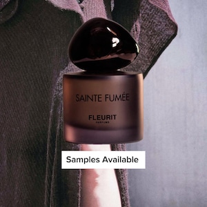 May include: A dark brown perfume bottle with a rounded top and a rectangular base. The bottle has the words "SAINTE FUMÉE" and "FLEURIT PARFUMS" printed on it. The text "Samples Available" is in a white rectangle below the bottle.