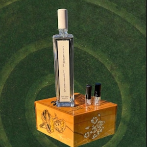 May include: A rectangular perfume bottle with a light-colored stopper and the text "SERGE LUTENS" in vertical lettering. The bottle sits on a decorative wooden box with floral designs, accompanied by two small vials. The background is a green, textured surface.
