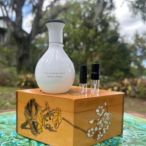 May include: A white perfume bottle with a black top and the text "The Harmonist Matrix Metal" sits on a wooden box with floral designs. Two small perfume vials with black caps are also on the box. The box is on a decorative table.