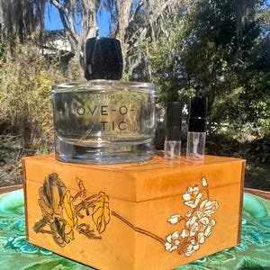 May include: A clear glass perfume bottle with a black cap and the text "LOVE-O-MATIC" sits atop a wooden box with floral designs. Two small sample vials are next to the perfume bottle. The box is on a green and gold surface.