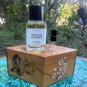 May include: A perfume bottle labeled "Vanilla Powder" and a small sample bottle sit on a decorative wooden box. The box has floral designs and is placed on a green and blue surface, with a natural background of trees and foliage.
