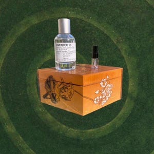 May include: A clear perfume bottle and a small vial sit atop a decorative wooden box with floral designs. The perfume bottle is labeled "ANOTHER 13" and contains 1.7 fl oz of perfume. The box is a warm orange color.