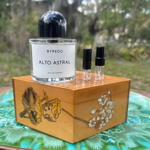 May include: A clear glass perfume bottle with a black cap sits atop a decorative wooden box. The bottle is labeled "BYREDO ALTO ASTRAL EAU DE PARFUM." Two small sample vials are next to the bottle. The box has floral designs.