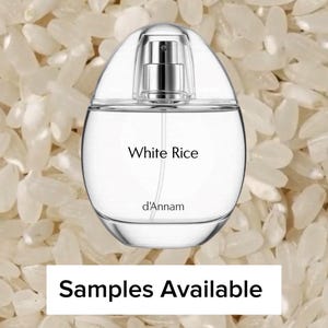 May include: A clear, oval-shaped perfume bottle with a silver atomizer, labeled "White Rice" by d'Annam. The bottle is set against a backdrop of white rice grains. The text "Samples Available" is displayed below.