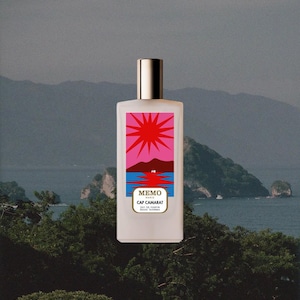 May include: A rectangular glass perfume bottle with a metallic cap. The label features a graphic illustration of a red sun over a dark mountain and blue water. The text on the label reads MEMO PARIS CAP CAMARAT EAU DE PARFUM. The bottle is opaque white.