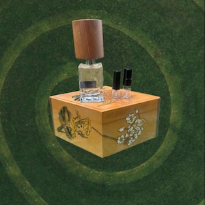 May include: A perfume bottle with a wooden cap and the text "Silver Musk" sits atop a decorative wooden box. Two small vials and floral artwork are also visible. The box is a warm yellow color.