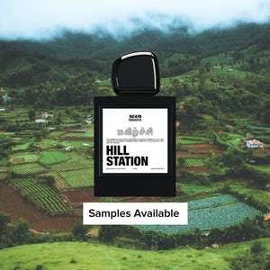 May include: A black perfume bottle with a square label featuring text in multiple languages, including "HILL STATION." The bottle has a glossy black cap. The background shows a green landscape with rolling hills and a cloudy sky. The text "Samples Available" is at the bottom.