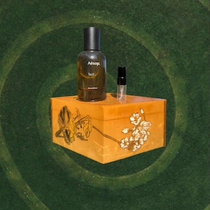 May include: A brown glass Aesop perfume bottle and a small vial sit atop a decorative wooden box. The box is painted with floral designs in yellow and white. The background is a green spiral pattern.