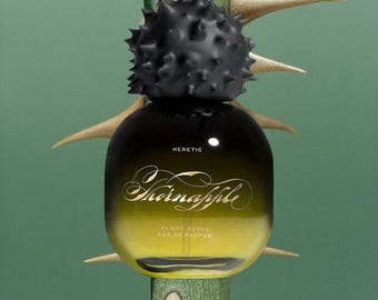 May include: A glass perfume bottle with a gradient yellow to black finish, featuring a black spiky cap. The bottle displays the text HERETIC, Thornapple, and PLANT-BASED EAU DE PARFUM. It is positioned in front of a green stem with sharp thorns.