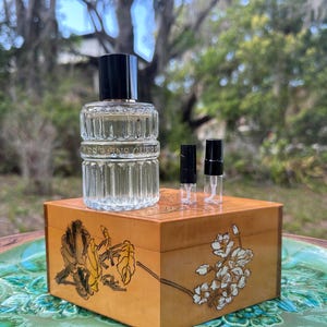 May include: A clear glass perfume bottle with a black cap and two small sample vials sit atop a wooden box with floral designs. The box is placed on a decorative green and brown ceramic plate, with a blurred outdoor background.