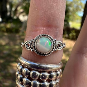 May include: A silver ring featuring a large, round opal gemstone with iridescent flashes of color. The opal is set in a twisted rope-like bezel, with decorative swirls on either side. The ring is shown on a finger, with other silver rings stacked below.