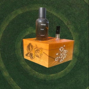 May include: A dark brown perfume bottle labeled "Aesop Karst" and a small sample bottle sit atop an orange wooden box with floral designs. The box is on a green background.