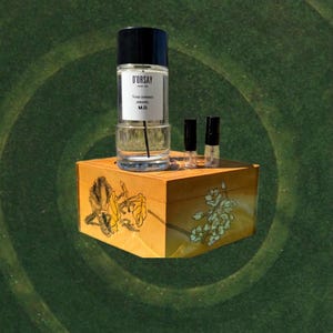 May include: A perfume bottle with a black cap and a label that reads "D'ORSAY". The bottle sits on a decorative wooden box with floral designs. Two small sample vials are also present. The background is a green, textured surface.