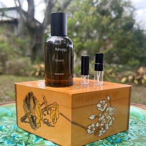 May include: A brown glass bottle of Aesop Gloam Eau de Parfum with a black cap, and two small sample vials, sit atop a decorative wooden box with floral designs. The box is placed on a green and blue patterned surface.