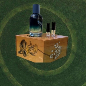 May include: A perfume bottle with a dark blue cap and a gradient blue-green liquid sits on a decorative wooden box. Two small vials are next to the bottle. The box is decorated with floral designs in yellow and white.
