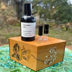 May include: A black perfume bottle with a white label, two small sample vials, and a decorative wooden box. The box is painted with floral designs in gold and black. The perfume bottle is labeled "PERFUMER H MOSS" and contains 50ml or 1.6 fl oz.