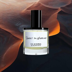 May include: A clear glass perfume bottle with a black cylindrical cap containing a pale yellow liquid. The white label features the black text Sweet Do Nothing, D.S. & DURGA, and eau de parfum 50 ml. The bottle has a thick base and straight sides.