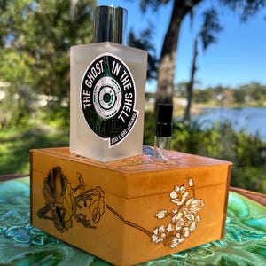 May include: A frosted glass perfume bottle with a silver cap and a label that reads "THE GHOST IN THE SHELL". The bottle sits on a wooden box with floral and insect designs. A small vial is next to the bottle.