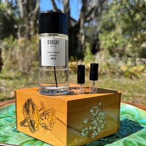 May include: A perfume bottle with a black cap and a white label that reads "D'ORSAY" and "Nous sommes amants M.D." sits on a wooden box with floral designs. Two small perfume vials with black caps are also present.