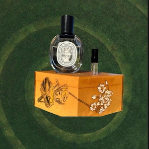 May include: A clear glass perfume bottle with a black cap and a small sample bottle sit atop a decorative orange box. The box is adorned with floral designs in white and yellow. The background is a green swirl.
