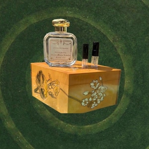 May include: A clear glass perfume bottle with a gold cap sits atop a wooden box with floral designs. Two small vials are next to the bottle. The box is a light brown color with yellow and white floral accents. The background is a dark green.