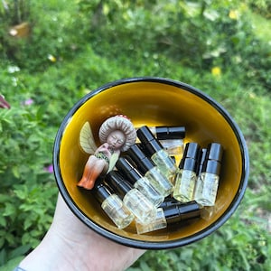 Puede incluir: Un cuenco redondo con exterior negro e interior amarillo dorado que contiene varios frascos peque&ntilde;os de vidrio transparente con tapones negros y una peque&ntilde;a figura de un hada con sombrero de seta y alas transl&uacute;cidas.