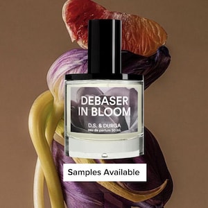 May include: A perfume bottle with a black cap and a white label that reads "DEBASER IN BLOOM". The bottle is surrounded by purple and yellow flowers and a slice of red fruit. The text "Samples Available" is at the bottom.