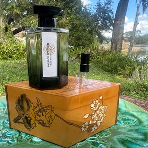 May include: A perfume bottle with a black cap and a white label that reads "PASSAGE D'ENFER EAU DE TOILETTE" sits on a wooden box with floral designs. A small, empty spray bottle is next to it. The box is on a green, patterned surface.