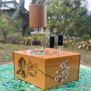 May include: A perfume bottle with a wooden cap and the text "Silver Musk" sits atop a decorative wooden box. Two small, empty perfume vials with black caps are next to the bottle. The box features floral and leaf designs in gold and black.