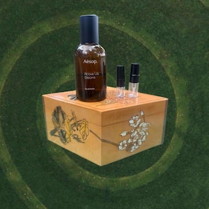 May include: A brown glass bottle of Aesop Above Us, Steorra Eau de Parfum with a dark blue cap, and two small sample vials, sits atop a decorative wooden box with floral designs. The box is light brown with painted flowers.