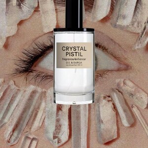 May include: A clear glass perfume bottle with a black cylindrical cap and a rectangular beige label. The label features the black text CRYSTAL PISTIL, fragrance/enhancer, D.S. & DURGA, and eau de parfum 100 ml. The bottle is centered over an eye.