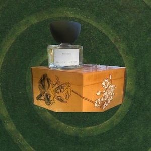 May include: A clear glass perfume bottle with a black cap sits atop a decorative wooden box. The bottle has a white label with the word "Noisette" and "MAISON D'ETTO". The box is light brown with floral designs.