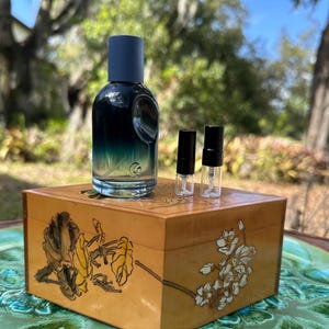May include: A clear glass perfume bottle with a dark blue cap and a gradient blue-green liquid inside. Two small sample vials sit next to the bottle on a wooden box with floral designs. The box rests on a green, patterned surface.
