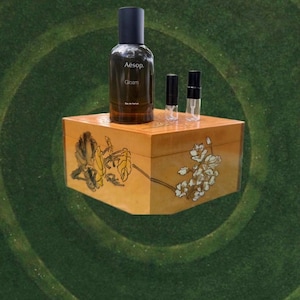 May include: A brown glass bottle of Aesop Gloam Eau de Parfum with two small sample vials on a decorative wooden box. The box is painted with floral designs in yellow and white.