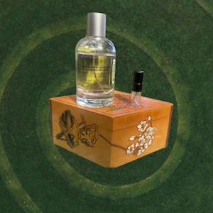 May include: A clear glass perfume bottle with a silver cap and a small sample bottle sit atop a wooden box. The box is decorated with floral designs in yellow and white. The background is a green, circular pattern.