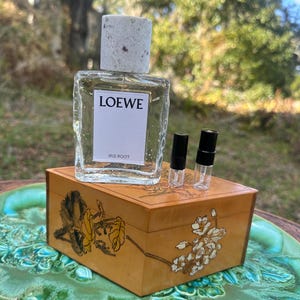 May include: A clear Loewe perfume bottle with a light gray cap sits atop a small, decorated wooden box. The box features floral designs in gold and brown. Two small, black-capped vials are also on the box. The perfume bottle has a white label that reads "LOEWE IRIS ROOT."