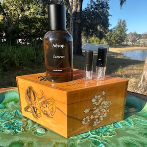 May include: A brown glass bottle of Aesop Ouranon Eau de Parfum with a black cap, and two small sample vials, sit on a decorative wooden box with floral designs. The box is on a green and blue decorative plate.