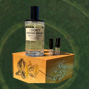 May include: A clear glass perfume bottle with a black cap and the text "I DON'T KNOW WHAT fragrance / enhancer" sits on a decorative wooden box. Two small sample vials are next to the bottle. The box has floral artwork.